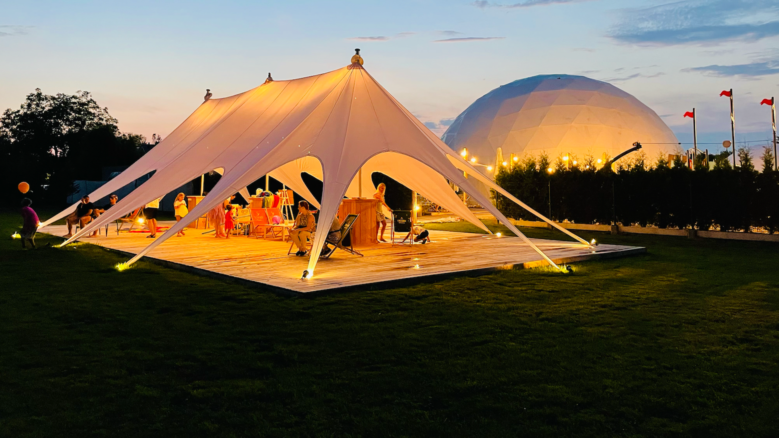 Illuminated stretch-tent lounge on a wooden deck with seating and guests at dusk, domed event hall and lawn in background