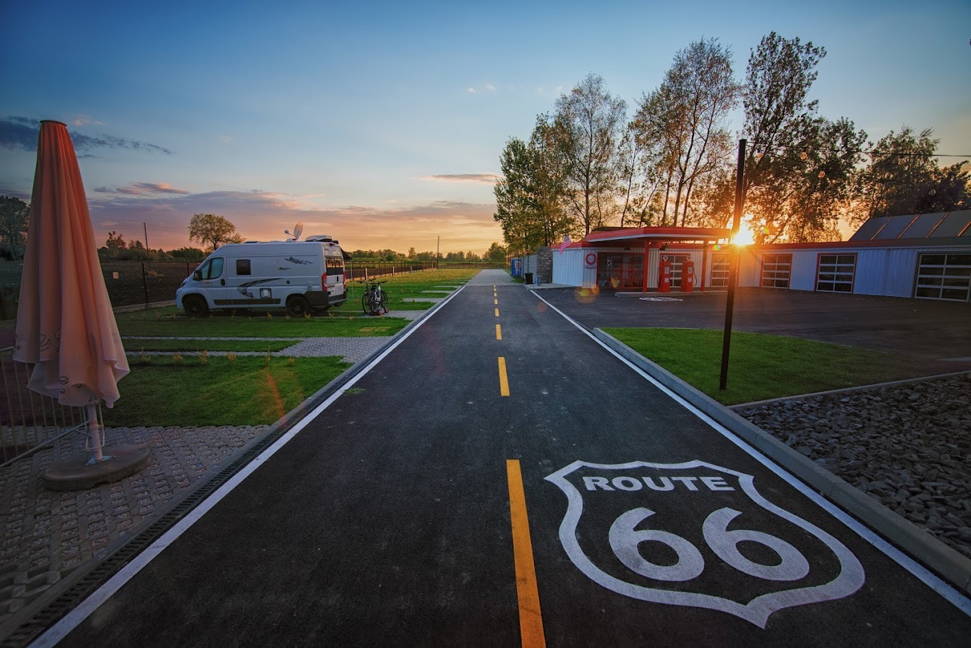 Campervan pitch beside a paved Route 66 lane at sunset, grassy pitches, service building and outdoor umbrella.