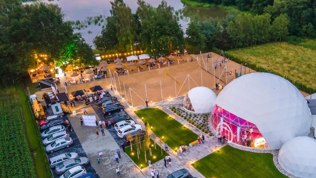 Lakeside evening event with illuminated geodesic domes glowing pink, sandy volleyball courts, lawn seating and guests