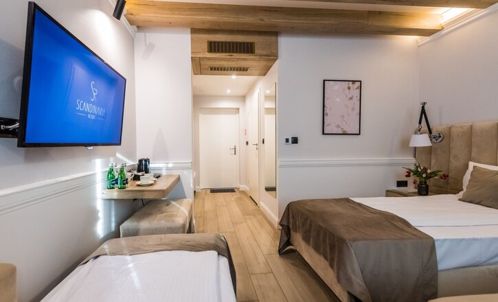 Bright modern hotel room with two beds, exposed wooden ceiling beams, wall TV, tea station with bottled water and bedside flowers.