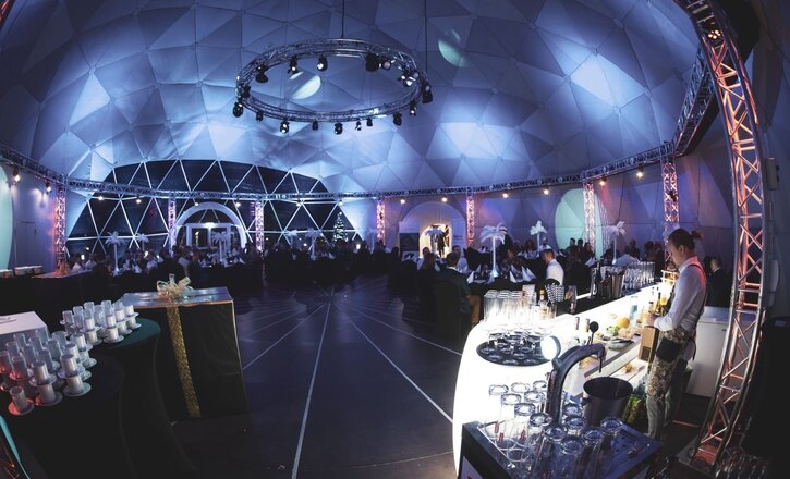 Spacious domed event hall with blue ambient lighting, circular truss of stage lights, seated dinner tables and bartender at illuminated bar.
