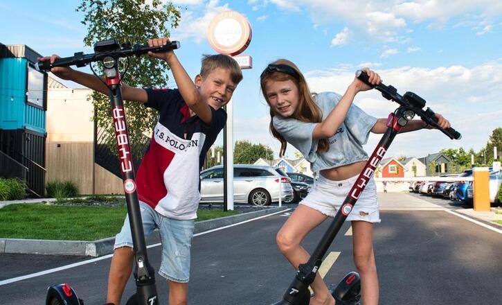 Two children riding electric scooters in a parking area with modern low-rise lodges and parked cars, smiling and leaning toward the camera.