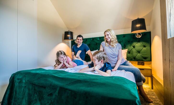 Family relaxing on a plush bed in a modern attic hotel room with green velvet headboard, bedside lamps and cozy lighting