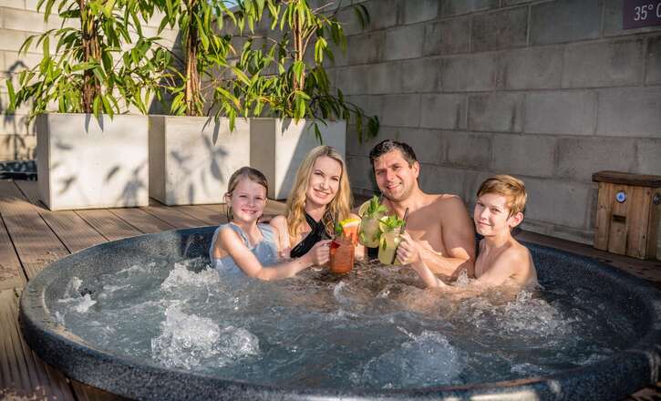 Family of four relaxing in an outdoor hot tub on a wooden terrace, clinking refreshing cocktails beside potted plants.
