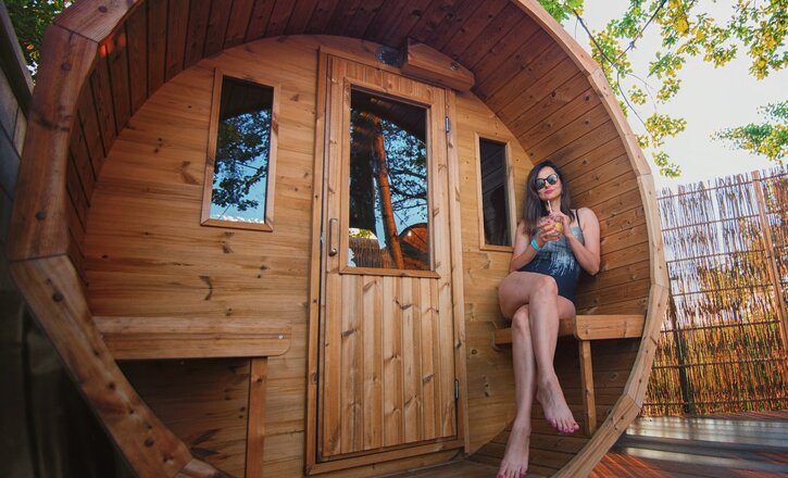 Barrel-shaped outdoor wooden sauna pod on private deck, woman in swimsuit relaxing with a drink on built-in bench under trees and string lights.