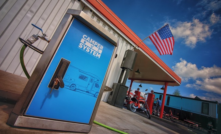 Blue "Camper System" RV water cabinet with hose at a covered service bay, parked motorcycles, picnic benches and a US flag