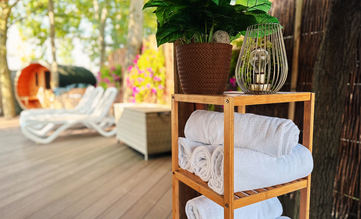 Rolled white towels on a wooden shelf with potted plant and decorative lantern, sun loungers and barrel sauna on shaded spa deck.