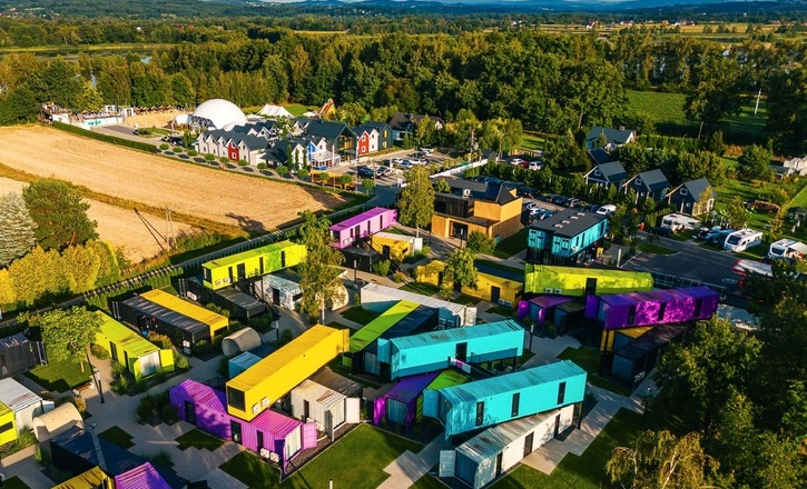 Kolorowy kompleks hotelowy z pokojami w kontenerach, trawnikami i alejkami, parkingiem, placem zabaw i wiejskimi widokami.
