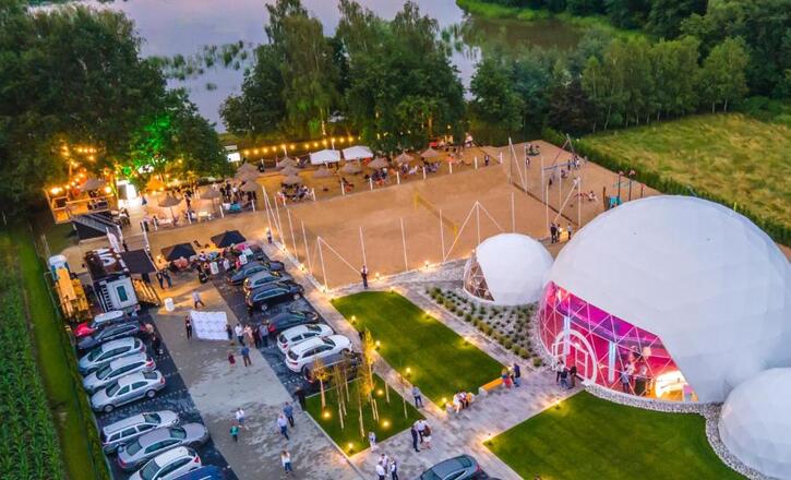 Lakeside evening event with illuminated geodesic domes glowing pink, sandy volleyball courts, lawn seating and guests