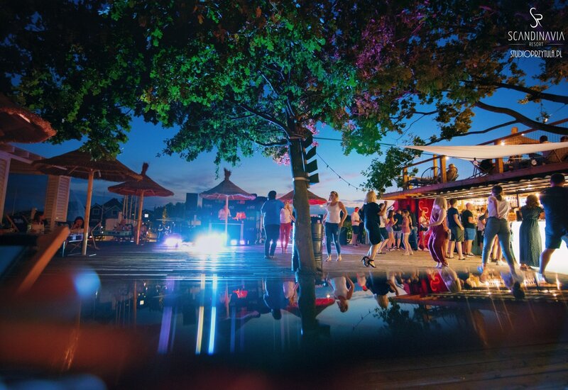 Wieczorny bar przy basenie w resorcie: goście tańczą pod podświetlonym drzewem, słomiane parasole, lampki i odbicia w wodzie.