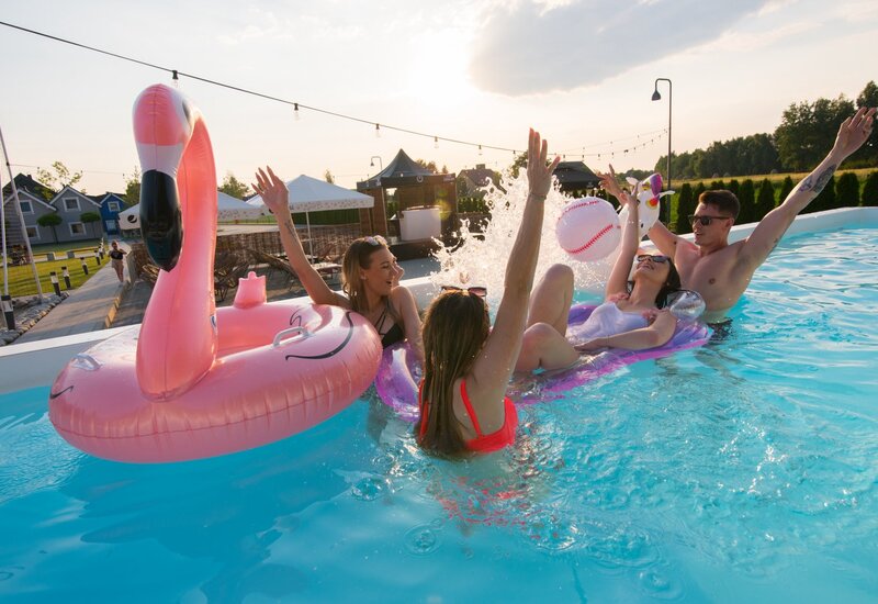 Goście bawią się w zewnętrznym basenie o zachodzie słońca, chlapiąc na różowym flamingu i jednorożcu przy altanach.
