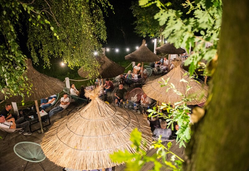 Hotelowy taras nocą z trzcinowymi parasolami, lampkami, wygodnymi fotelami i gośćmi relaksującymi się przy stolikach