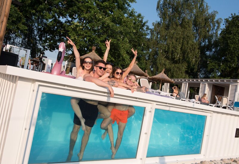 Grupa młodych gości pochylona nad basenem z przezroczystą ścianą, śmieją się, nogi widoczne przez szybę, leżaki i parasole w tle.