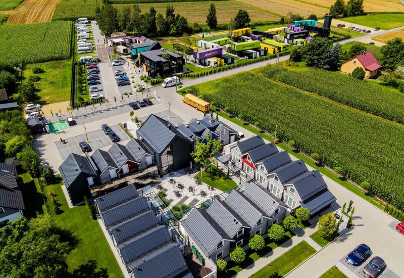 Nowoczesny ośrodek z lotu ptaka: rzędy domków z szarymi dachami, patio zewnętrzne z miejscami do siedzenia, parking i kolorowe moduły przy polach kukurydzy