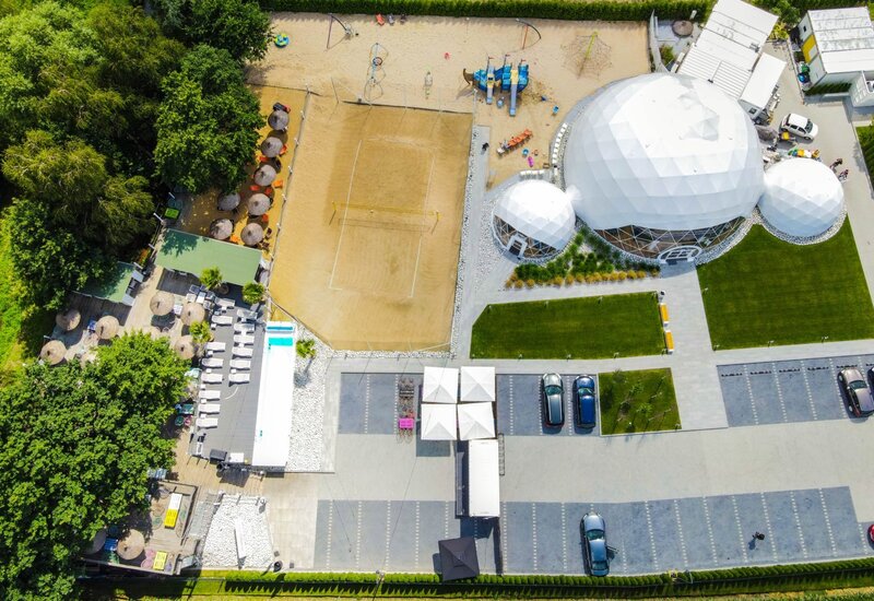 Biała kopuła geodezyjna jako przestrzeń eventowa przy piaszczystym boisku do siatkówki plażowej, taras z leżakami i parasolami, plac zabaw i parking