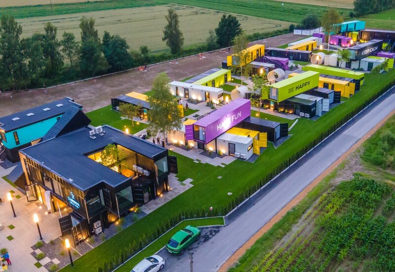 Kolorowy hotel z kontenerów z podświetloną recepcją, pokojami z prywatnymi tarasami, trawnikiem i parkingiem przy polach o zmierzchu