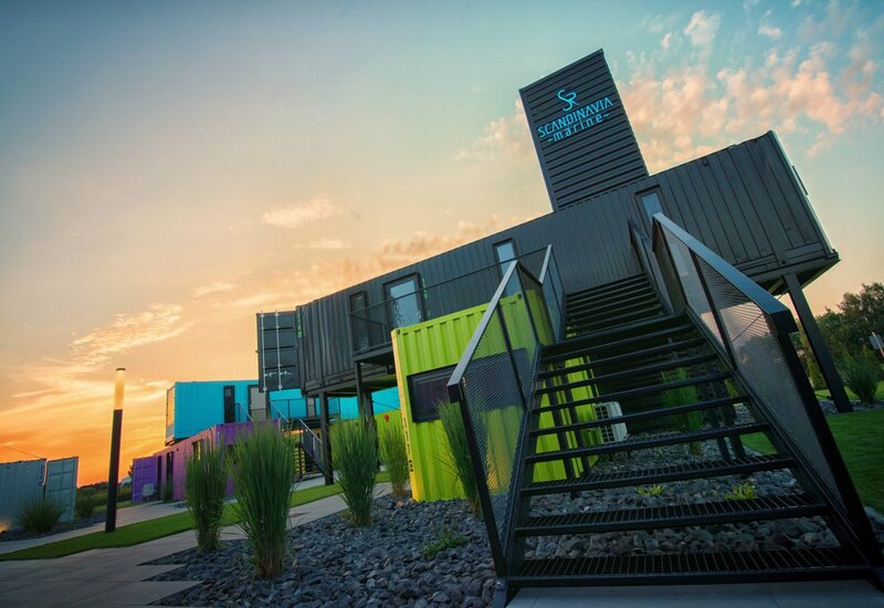 Kolorowy hotel z kontenerów z metalowymi schodami, kamienne rabaty i trawy przy zachodzie słońca pod napisem 'Scandinavia Marine'