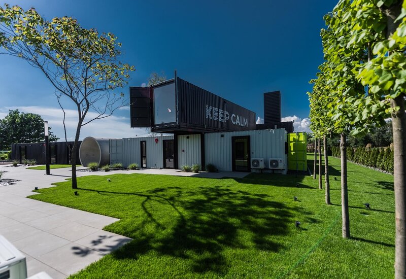Budynek z nałożonych kontenerów z napisem „KEEP CALM”, taras z płyt, zadbany trawnik, młode drzewa i zewnętrzne klimatyzatory