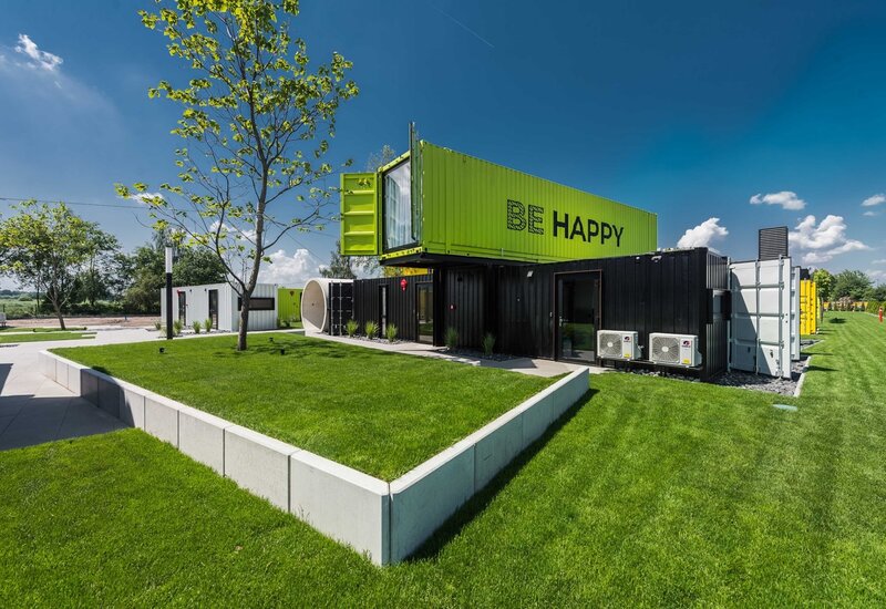 Modułowy hotel z kontenerów: jaskrawozielony pawilon „BE HAPPY” na czarnych modułach, zadbany trawnik i dziedziniec pod błękitnym niebem