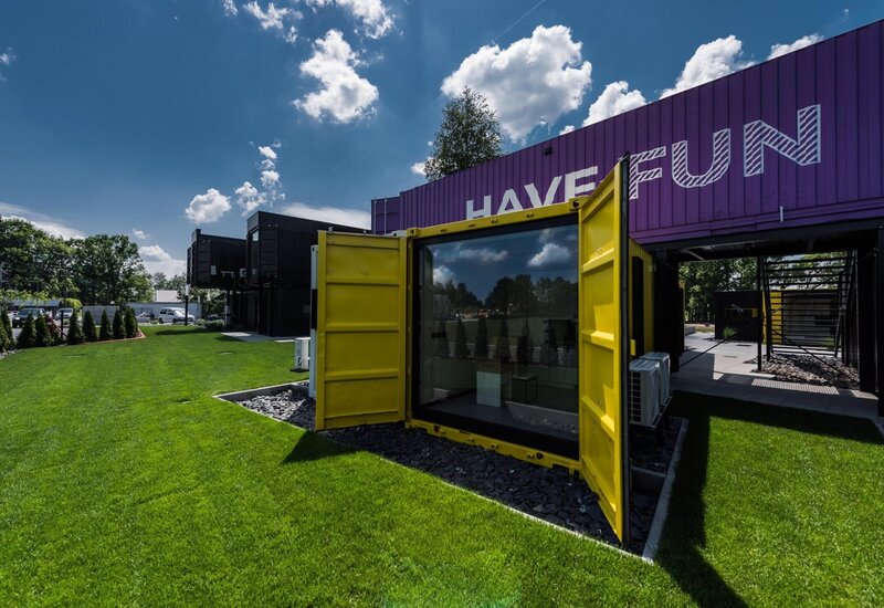 Modułowy hotel z kontenerów, żółwy moduł-pokój z otwartymi drzwiami i przeszkloną ścianą, fioletowa elewacja HAVE FUN nad trawnikiem