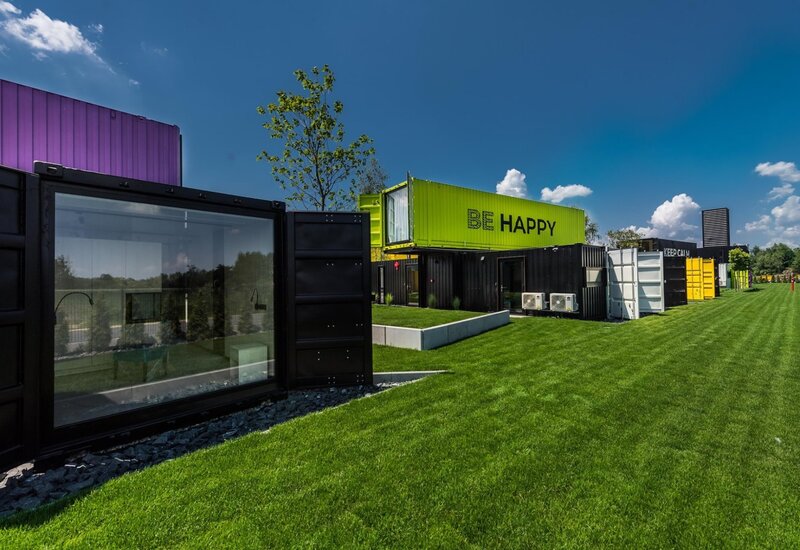 Kolorowy hotel z kontenerów z napisem 'BE HAPPY', szklanym pokojem i zadbaną trawą pod jasnym niebem