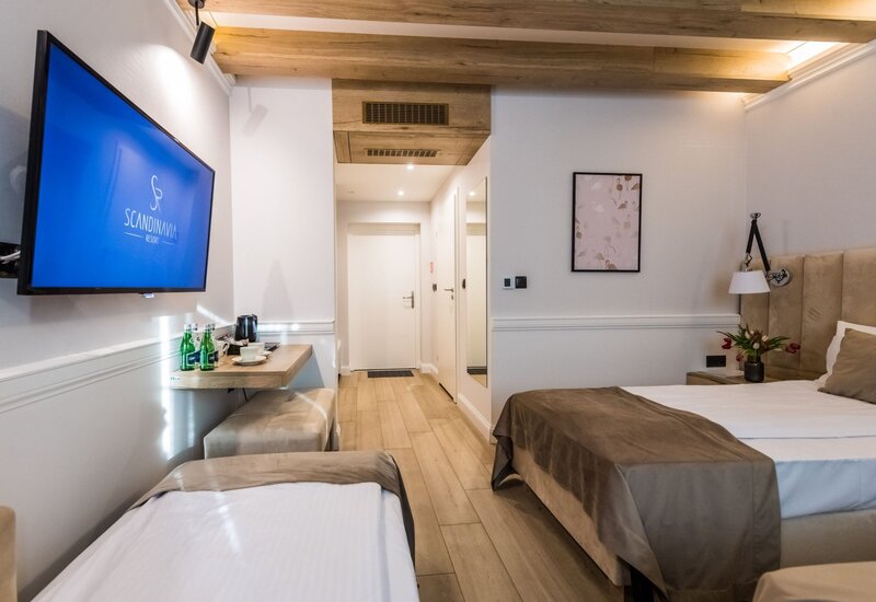 Jasny, nowoczesny pokój hotelowy z dwoma łóżkami, odkrytymi drewnianymi belkami, telewizorem, czajnikiem i butelkowaną wodą.