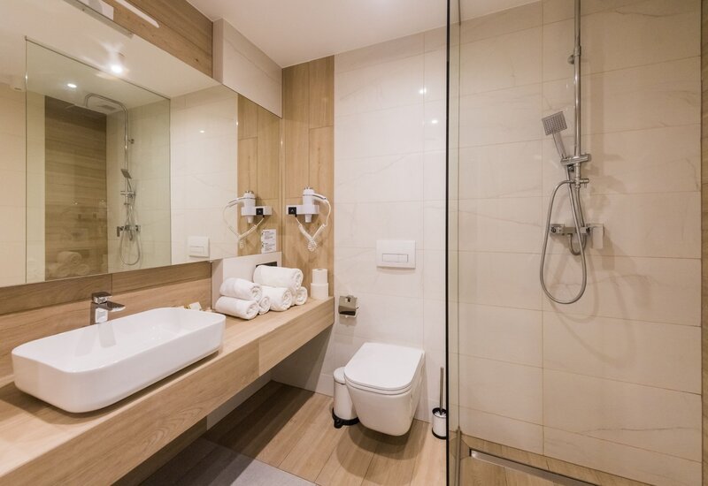 Nowoczesna łazienka hotelowa z drewnianą półką, dużą prostokątną umywalką, zwiniętymi ręcznikami, suszarkami i prysznicem z deszczownicą