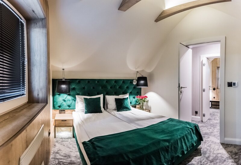 Butikowa sypialnia z butelkowozielonym pikowanym zagłówkiem, połączonymi łóżkami, lampkami nocnymi, sklepieniem z drewnianymi belkami i świeżymi kwiatami.