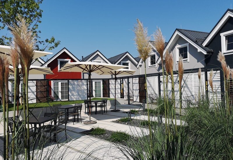 Słoneczne patio z parasolami, stolikami, ozdobnymi trawami i nowoczesnymi czerwono‑szarymi domkami z prywatnymi tarasami.