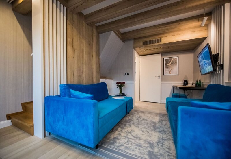 Przytulny salon apartamentu z intensywnie niebieskimi sofami, drewnianymi belkami sufitowymi, telewizorem, biurkiem i wazonem czerwonych róż.