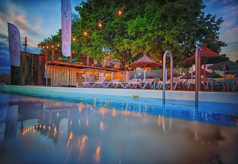 Bar przy basenie o zmierzchu z girlandami świateł, leżakami i słomianymi parasolami na drewnianym tarasie pod drzewami, odbicia w wodzie