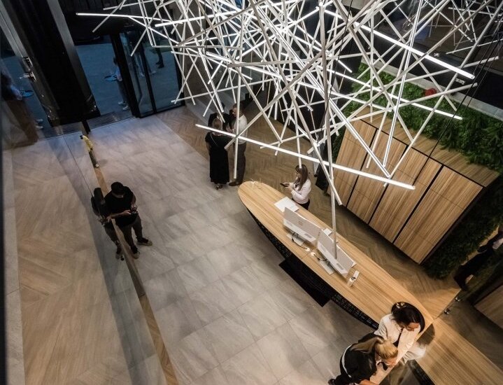 Nowoczesne lobby z geometryczną instalacją świetlną nad zaokrągloną recepcją, marmurową podłogą, zielenią i gośćmi.