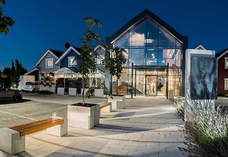 Szklane wejście do hotelu z podświetlonym atrium o zmierzchu, taras z parasolami, ławki, parking i nowoczesna zieleń.