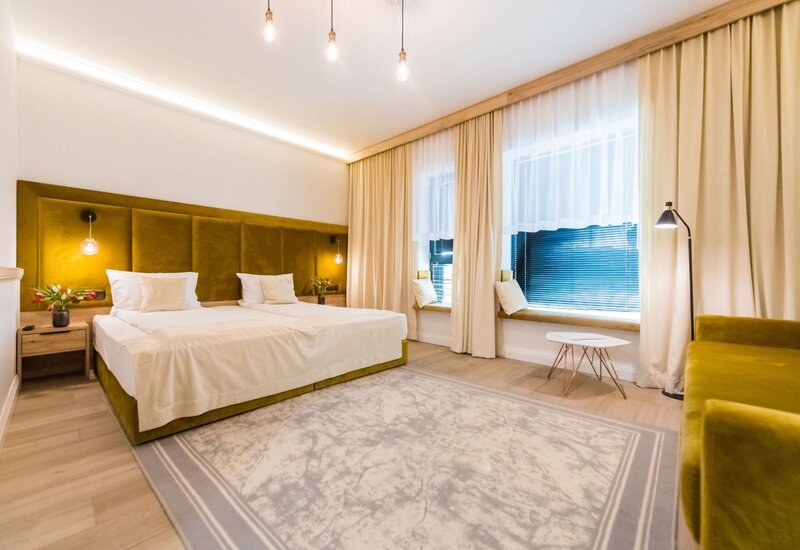 Nowoczesny pokój hotelowy z oliwkowym welurowym zagłówkiem, dwoma łóżkami, lampami wiszącymi, siedziskiem przy oknie i sofą