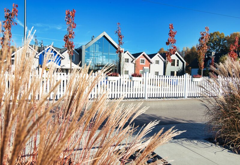 Butikowy hotel z przeszklonym lobby i kolorowymi domkami za białym płotem, ozdobne trawy i jesienne drzewa.