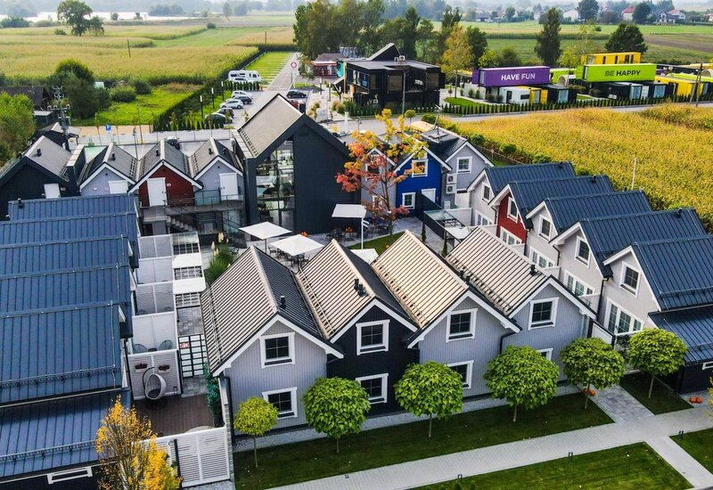 Butikowy ośrodek z kolorowymi domkami ze stromymi dachami, przeszklonym budynkiem wspólnym, tarasami z parasolami i widokiem na pola.