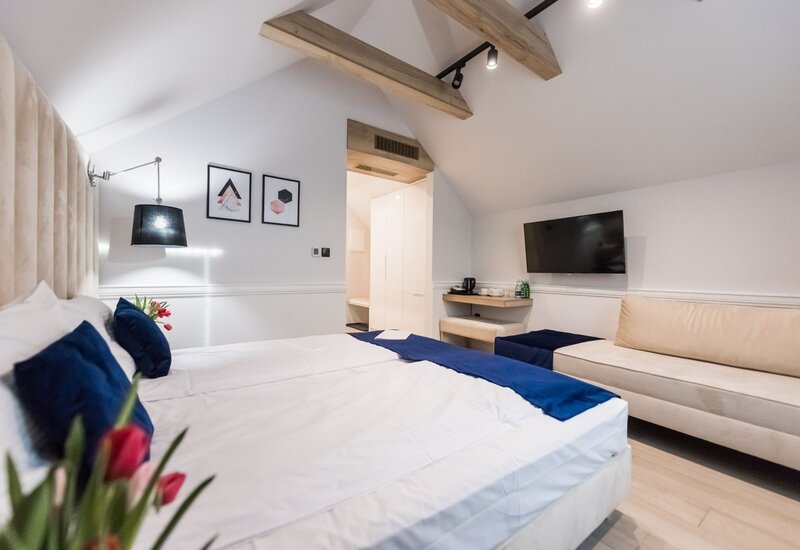 Poddasze — przytulny pokój hotelowy z pochylonym sufitem i drewnianymi belkami, dużym łóżkiem, granatowymi poduszkami, sofą, TV i czajnikiem