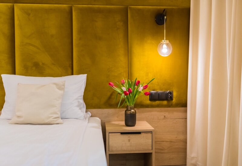 Łóżko z miękkim musztardowym welurowym zagłówkiem, białą pościelą, stolikiem nocnym z wazonem tulipanów i wiszącą szklaną lampą