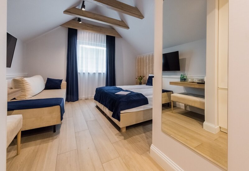 Strychowy pokój dwuosobowy z drewnianymi belkami, beżowym łóżkiem i sofą, granatowymi narzutami, TV i biurkiem z butelkami wody.