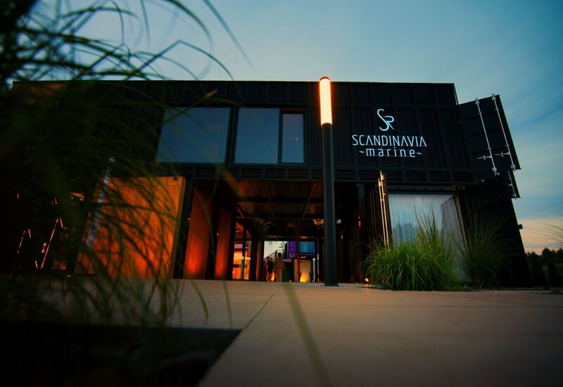 Wejście butikowego hotelu w czarnej kontenerowej bryle, neon 'Scandinavia Marine', oświetlony podjazd i dwaj goście o zmierzchu
