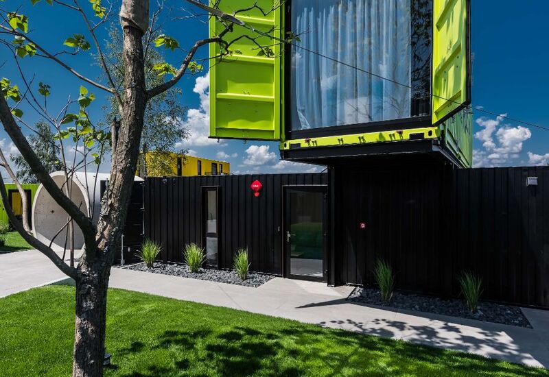 Podwieszany limonkowo-zielony apartament z kontenera z dużym oknem i zasłonami nad czarnymi pokojami na parterze, trawiasty dziedziniec