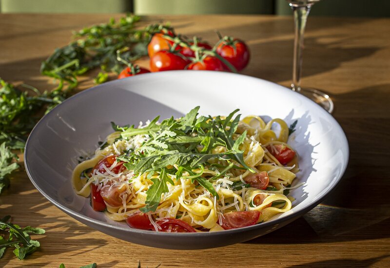 Tagliatelle z pomidorkami koktajlowymi, rukolą i parmezanem w białej misce, gałązka pomidorów i kieliszek białego wina na słonecznym stole