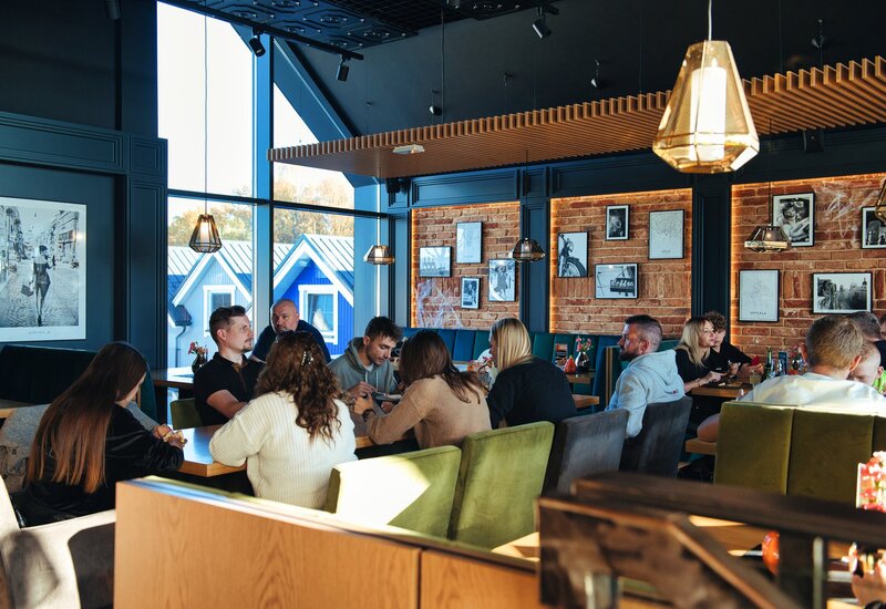 Przytulna restauracja hotelowa z ceglaną ścianą, drewnianym lamelowym sufitem, wiszącymi lampami, dużymi oknami, zielonymi aksamitnymi sofami i gośćmi przy posiłkach.
