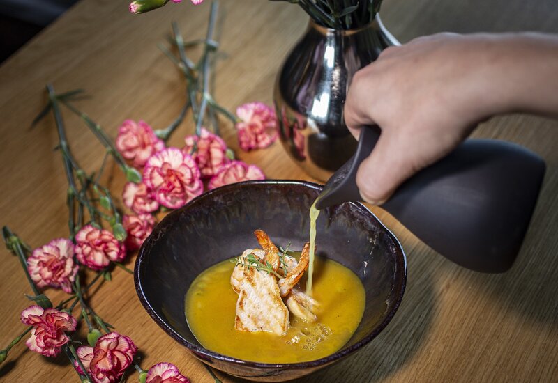 Elegancka miska z podsmażonymi owocami morza i krewetkami w złotym bulionie, dłoń dolewa sos; różowe goździki w wazonie na drewnianym stole.