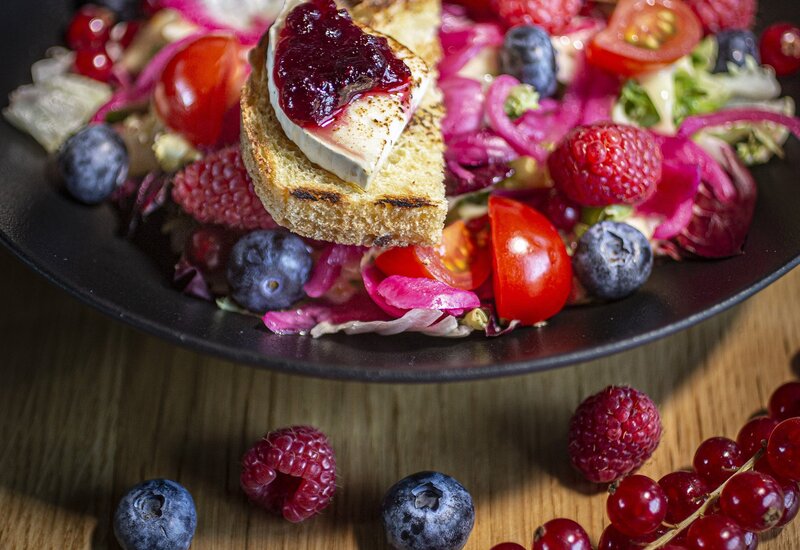 Grillowany kozi ser na crostini z dżemem jagodowym na mieszanej sałatce, maliny i borówki rozsypane na drewnianym stole