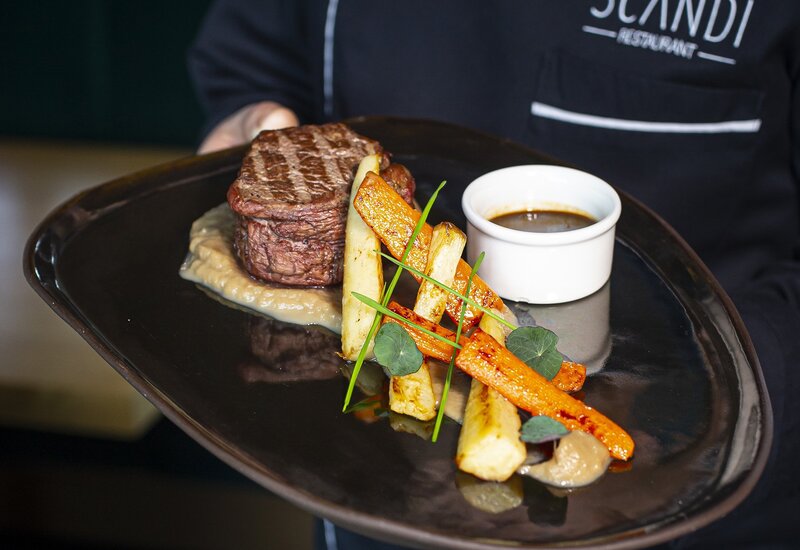 Kelner podaje grillowaną polędwicę na purée z pieczonymi warzywami korzeniowymi, mikrozieleniną i sosem na czarnym talerzu.