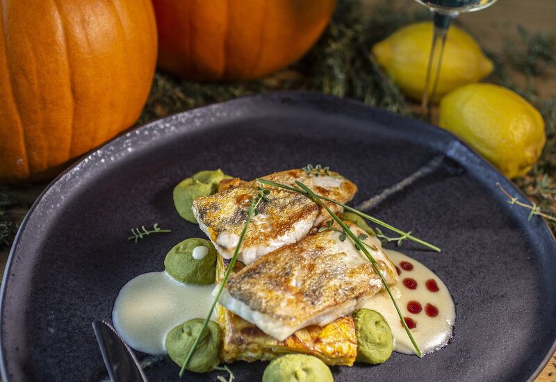 Smażona ryba na terrinie z dyni z puree z groszku, kremowym sosem i ziołami na ciemnym talerzu; kieliszek wina, cytryny i dynie