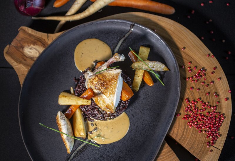 Pieczony kurczak na czarnym ryżu z glazurowanymi marchewkami, pasternakami i kremowym sosem na czarnym talerzu na drewnianej desce z różowym pieprzem