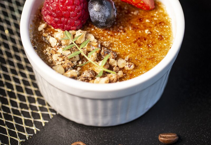 Krem brulee w białej kokilce ze skarmelizowaną skorupką, maliną, borówką, połówką truskawki, kruszonymi orzechami i tymiankiem; ziarna kawy obok