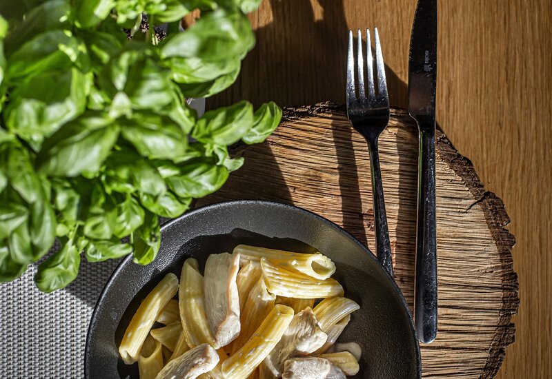 Kremowe penne z kurczakiem w czarnej misce, świeża bazylia obok, nóż i widelec na rustykalnej drewnianej podstawce, słoneczne nakrycie stołu.